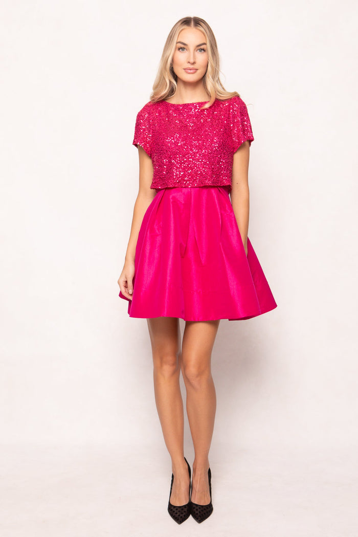 A woman stands against a white background wearing the Alexis Dress in Fuchsia by Ivy City Co—an eye-catching, bright pink sequin party dress with short sleeves and a matching flared skirt. She has long blonde hair and completes the look with black heels, making a bold statement for any special occasion.