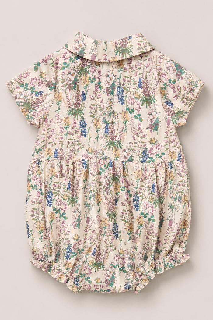 The Baby Amber Romper in Autumn Bouquet by Ivy City Co is a short-sleeved viscose romper showcasing a charming floral print in shades of purple, blue, and green on a cream background. Designed with a gathered waist, delicate lace trim, and buttons down the back, this airy and comfortable romper is perfect for your little one.