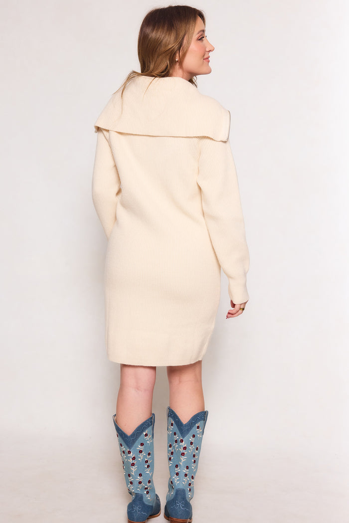 A woman faces away, wearing the Ivy City Co Briar Dress in Cream—a long-sleeve midi with a wide collar. Her light brown hair and blue cowboy boots with decorative sides complete the look against a plain white background.