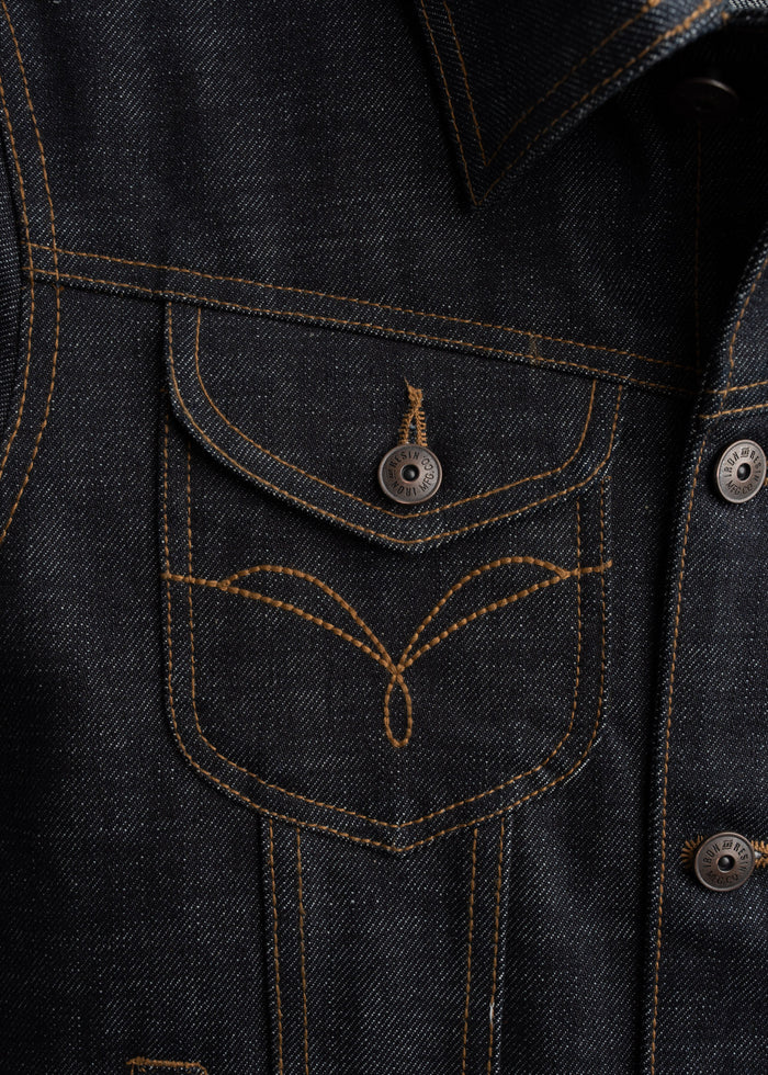 Close-up of a denim jacket pocket with orange stitching and metal buttons, showcasing detailed craftsmanship and texture.