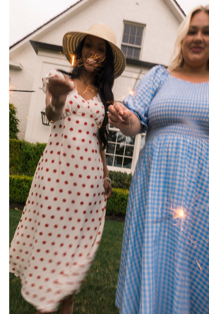 Two women in summery dresses, one wearing the Ivy City Co Fiona Dress in Red Polka Dot and the other in a blue gingham midi dress, stand outside a house holding sparklers, smiling and celebrating.