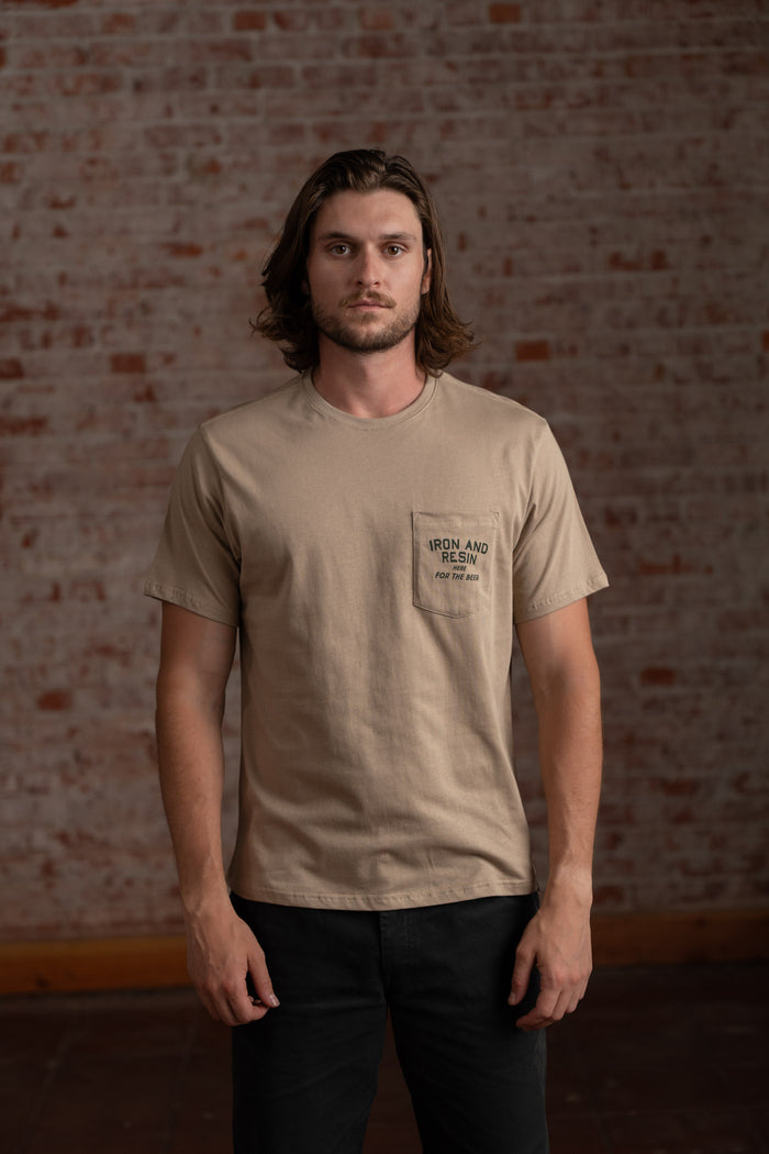 Man in beige "Iron and Resin" shirt stands against a brick wall, looking directly at the camera with a neutral expression.