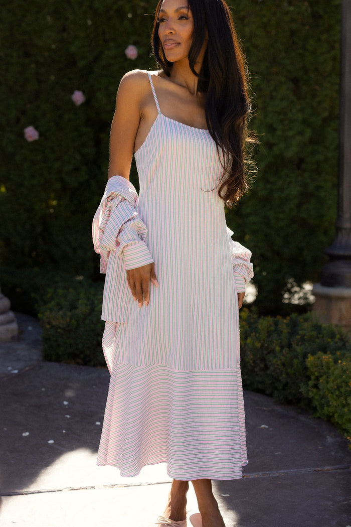 A woman stands outdoors among green bushes and pink flowers, wearing the Marbella Dress in Green & Pink Pinstripe by Ivy City Co— a sleeveless midi dress with thin straps and light pastel stripes — while holding a matching long-sleeve top.