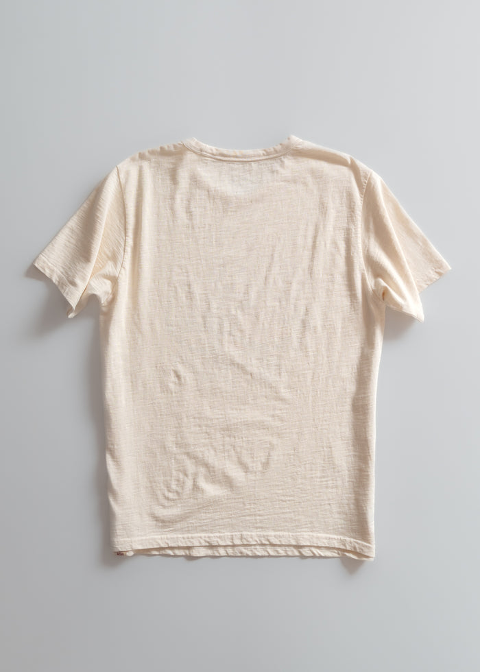 A plain beige T-shirt is laid flat on a light gray background, showcasing its texture and short sleeves in a minimalist style.