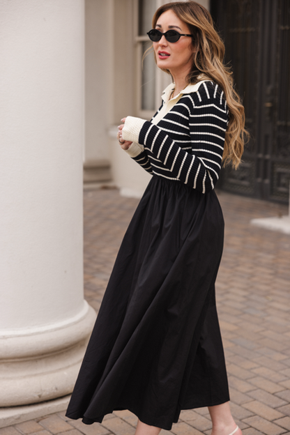 A woman in sunglasses walks outdoors on a brick path near white columns and a dark ornate door, wearing the Ivy City Co Adelina Dress in Black featuring a striped knit bodice.