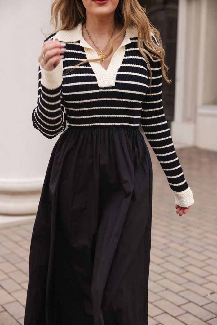 A woman with wavy hair wears the Adelina Dress in Black by Ivy City Co, featuring black and white stripes with a cream collar and cuffs. She holds a necklace while standing on a tiled outdoor walkway.
