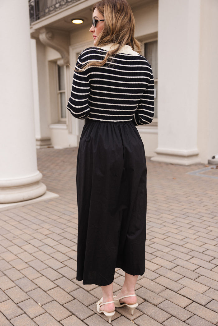 A woman with long, wavy hair stands on a brick walkway facing away, wearing sunglasses and the Adelina Dress in Black by Ivy City Co. Cream-colored heeled sandals complete her look against white columns and a building.