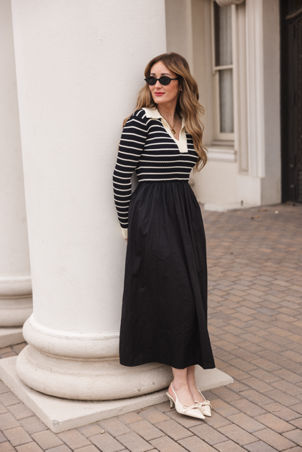 A woman with wavy hair and sunglasses stands by a white column on a brick walkway, wearing the Ivy City Co Adelina Dress in Black and cream pointed bow shoes.