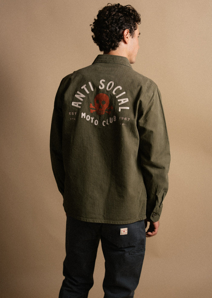Man in olive jacket with "Anti Social Moto Club" and skull design on back, stands against a beige background, facing away.