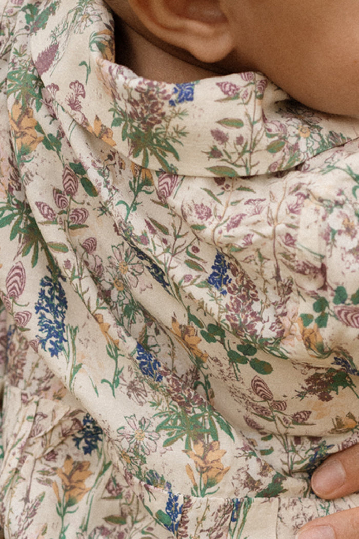 A close-up of a child's shoulder and neck wearing the Baby Amber Romper in Autumn Bouquet by Ivy City Co—a cream-colored romper adorned with a vibrant floral pattern in green, purple, blue, and yellow, elegantly finished with delicate lace trim.