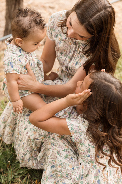 A woman and two children, all dressed in matching floral dresses adorned with delicate lace trim, sit together outdoors. The woman cradles a baby who’s wearing the Ivy City Co Baby Amber Romper in Autumn Bouquet, while an older child sits nearby, smiling and gently touching her face.