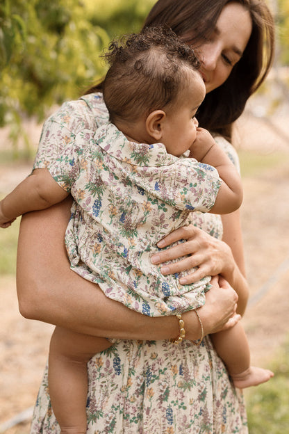 A woman in a floral dress holds a baby dressed in the Ivy City Co Baby Amber Romper in Autumn Bouquet, featuring delicate lace trim, while outdoors. Both look content, with the woman’s face partially visible and the baby resting comfortably in her arms.
