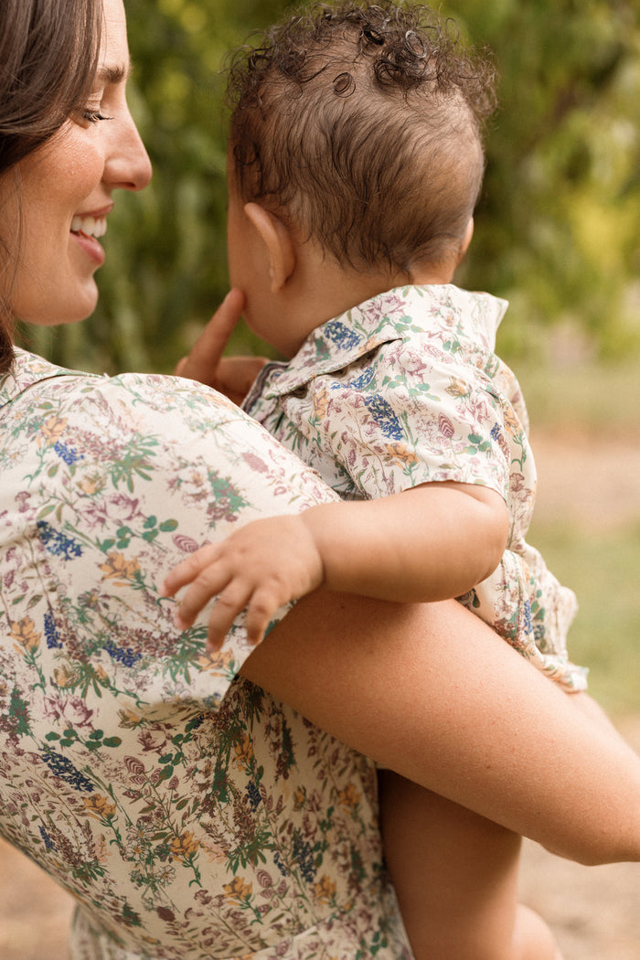 A woman stands outdoors, smiling at a baby in her arms as greenery blurs in the background. Both wear matching floral-patterned outfits, with the baby dressed in Ivy City Co’s Baby Amber Romper in Autumn Bouquet, beautifully accented by delicate lace trim. The baby faces away and gently touches the woman’s lips.