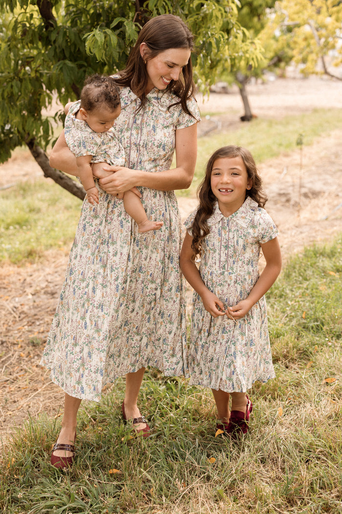 A woman and two young girls, all dressed in matching floral outfits with delicate lace trim, stand outside on the grass near trees. One girl is held by the woman, while the other—wearing the Ivy City Co Baby Amber Romper in Autumn Bouquet—stands smiling beside them.