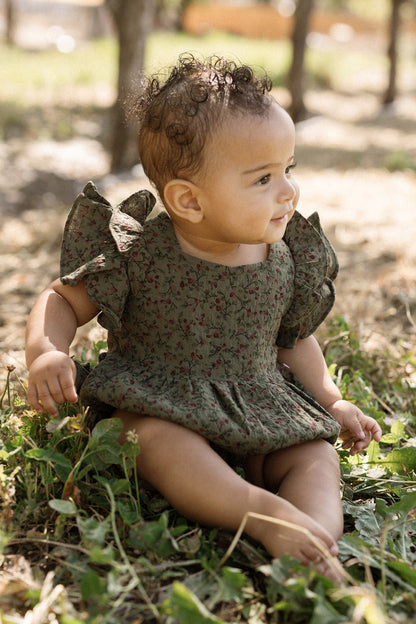A baby sits on the grass outdoors, smiling in the sunlight and looking to the side while wearing the Ivy City Co Baby Hattie Romper in Woodland Floral with flutter sleeves.