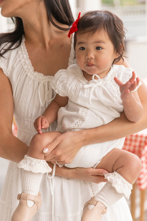 A woman in white holds her baby girl dressed in the Ivy City Co Baby Palermo Romper in White, accessorized with frilly socks, beige shoes, and a red bow—the perfect matching moment for mom and daughter.