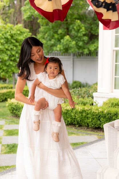 A woman in a white dress holds a baby girl wearing the Ivy City Co Baby Palermo Romper in White with a red bow, outside on a patio with greenery and an American flag banner, both enjoying their matching Mom and Me outfits.