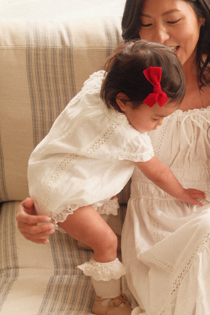 A woman in a white dress smiles at a toddler wearing the Ivy City Co Baby Palermo Romper in White with a red bow, as the child climbs onto her lap for a sweet Mom and Me matching moment.