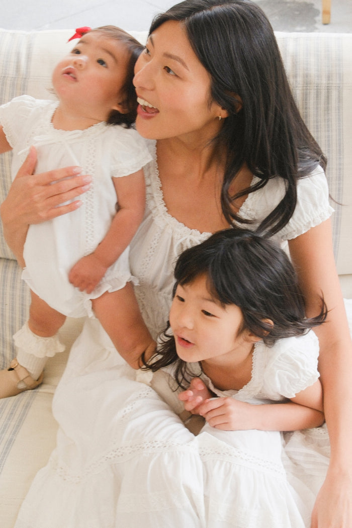A woman in a white dress sits on a couch, holding a baby in the Ivy City Co Baby Palermo Romper in White. A young girl, also dressed in white, leans against her. All three with dark hair enjoy their matching Ivy City Co “Mom and Me” outfits.