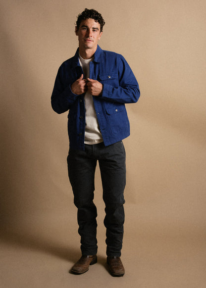 Man in blue jacket, white shirt, dark pants, and brown boots stands against a beige background, hands adjusting jacket.
