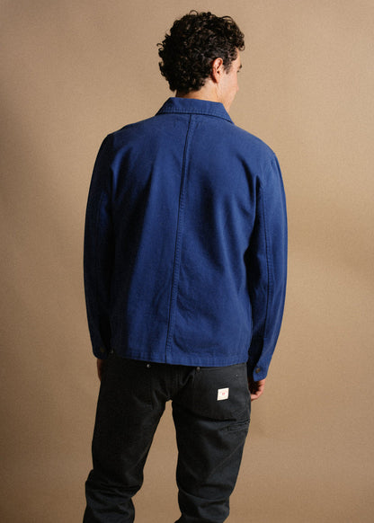 Man in blue jacket and black jeans stands with back to camera against a beige background, showcasing casual fashion style.