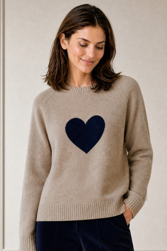 Model wearing the Aspiga Cashmere Crew Neck Heart Jumper in Oat/Navy, a crew-neck jumper with a contrast heart motif, over navy trousers, front view