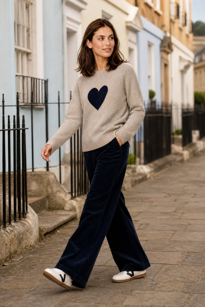 Model wearing the Aspiga Cashmere Crew Neck Heart Jumper in Oat/Navy with navy corduroy trousers, walking outdoors on a street