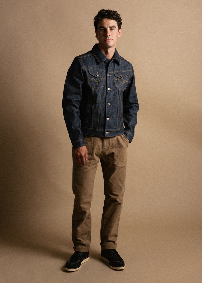 Man in a denim jacket and khaki pants stands against a beige background, exuding a casual yet stylish look.