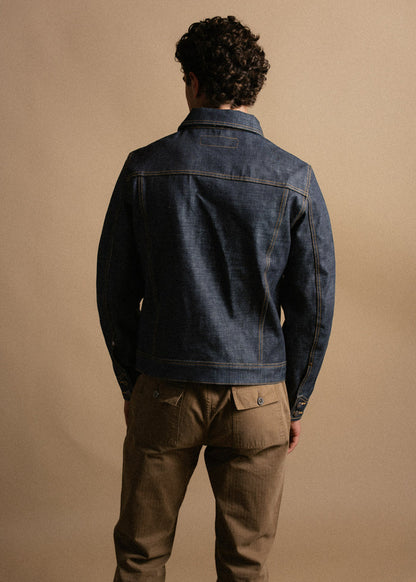 Man in denim jacket and brown pants stands with back to camera against a beige background.