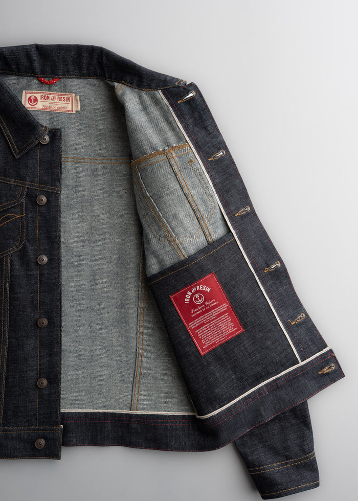Open denim jacket showing Iron & Resin label inside. Red patch with text on the inner pocket. Gray background.