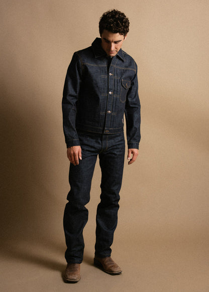 Man in dark denim jacket and jeans stands against a beige background, looking down, wearing brown boots.