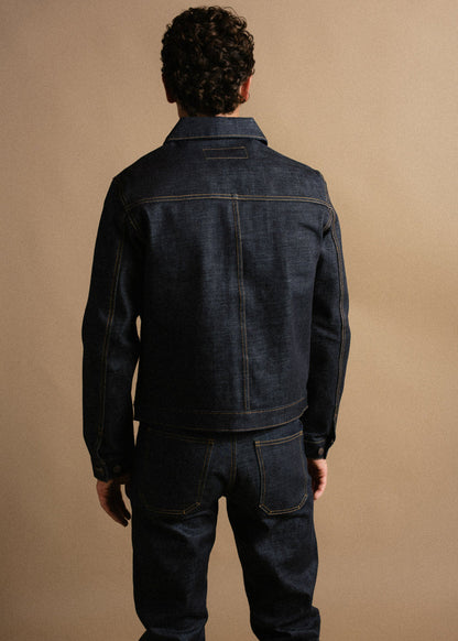 Man in dark denim jacket and jeans stands with back to camera against a beige background.
