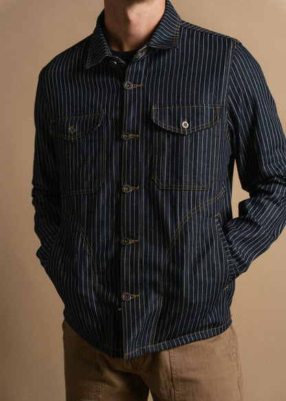 Man in a striped denim jacket with buttoned pockets and brown pants stands against a beige background, hands in pockets.