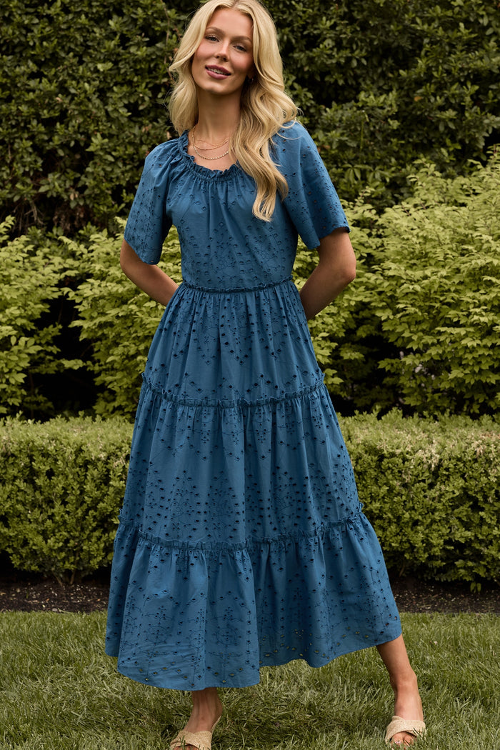 A woman smiles outdoors in front of green bushes, wearing Ivy City Co’s Etta Dress in Cobalt—a summer midi with a gathered neckline, subtle dotted pattern, short sleeves, side pockets, and her long blonde hair enhances the breezy look.