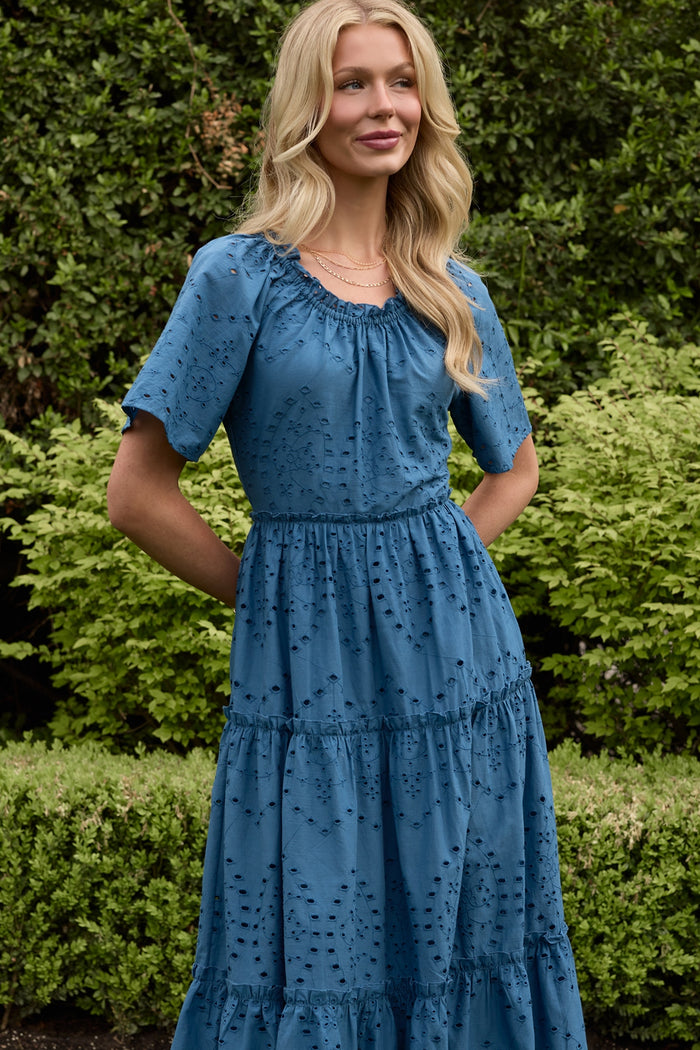 A woman with long blonde hair smiles outdoors in front of greenery, hands in the pockets of her Ivy City Co Etta Dress in Cobalt.