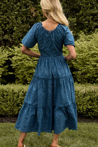 A woman with long blonde hair stands outdoors, her back to the camera, wearing the Etta Dress in Cobalt by Ivy City Co—a summer midi dress with a tiered skirt and embroidered details—set against lush green foliage.