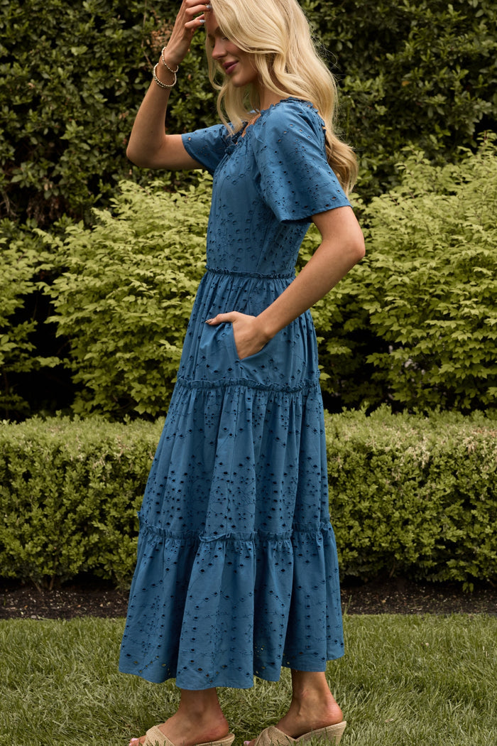 A woman with long blonde hair smiles while wearing the Ivy City Co Etta Dress in Cobalt. She stands outdoors on grass with greenery in the background, facing sideways with one hand in her dress pocket.