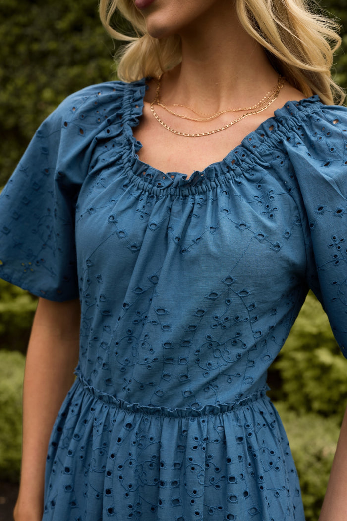A woman wears the Ivy City Co Etta Dress in Cobalt, a summer midi with puffed short sleeves and a ruffled neckline. She accessorizes with layered gold necklaces; her blonde hair is loose and greenery is blurred in the background.