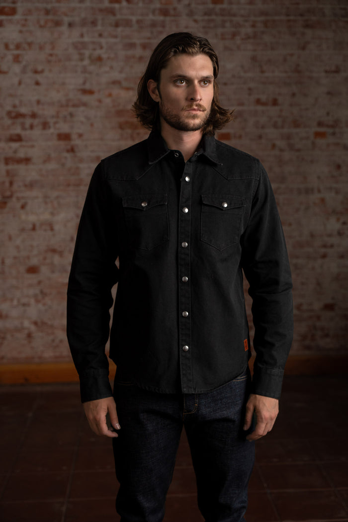 Man in a black button-up shirt stands against a rustic brick wall, exuding a casual yet confident style in a dimly lit room.