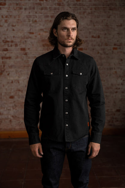 Man in a black button-up shirt stands against a rustic brick wall, exuding a casual yet confident style in a dimly lit room.