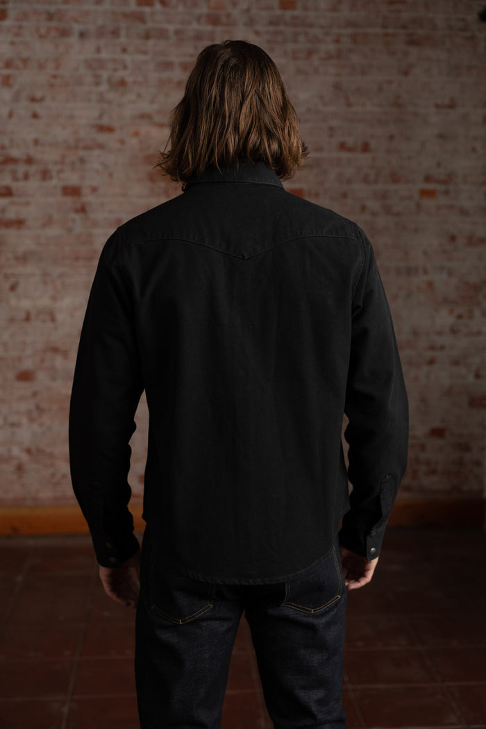 A person with long hair stands facing a brick wall, wearing a black shirt and dark jeans, creating a casual, relaxed vibe.