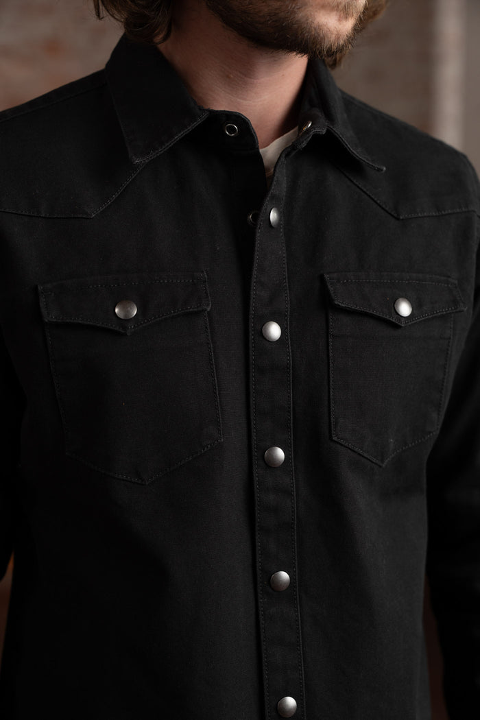 Man in a black denim shirt with silver buttons and chest pockets, standing against a blurred brick wall background.
