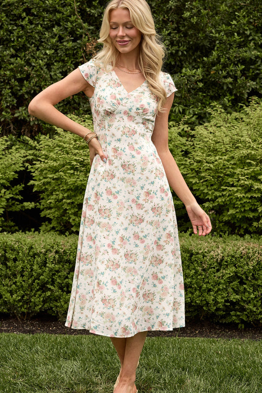 A woman smiles outdoors before green bushes, adjusting her hair in the Ivy City Co Fiona Dress in Countryside Peony Blossom—a light, short-sleeved summer dress with a delicate floral print.