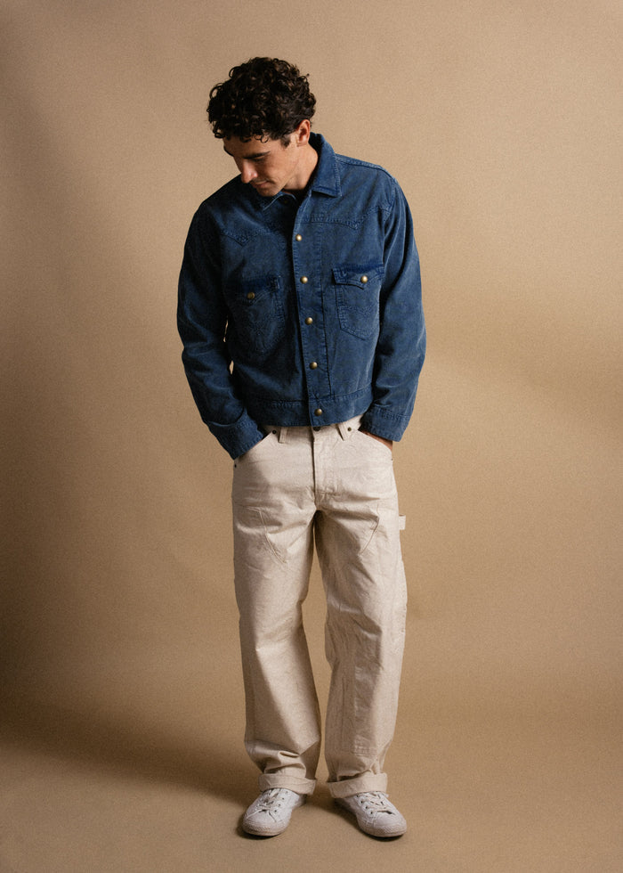 Man in blue denim jacket and beige pants looks down, standing against a plain tan background, hands in pockets, casual style.