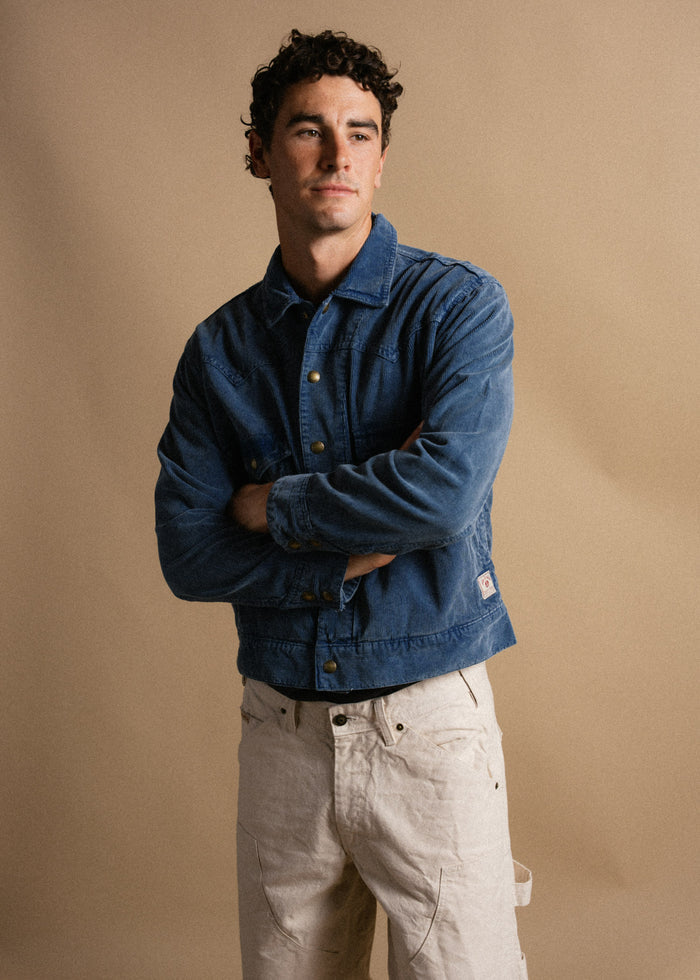 Man in blue denim jacket and beige pants stands against a neutral background, arms crossed, looking thoughtfully to the side.