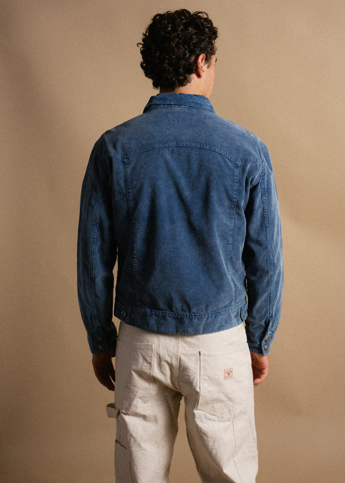 Man in blue denim jacket and white pants stands against a beige background, facing away, highlighting casual fashion style.