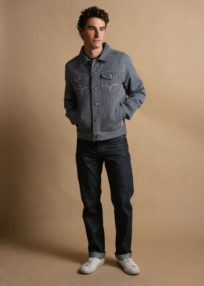 Man in striped jacket and jeans stands against a beige background, hands in pockets, looking to the side, wearing white sneakers.