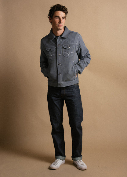 Man in striped jacket and jeans stands against a beige background, hands in pockets, looking to the side, wearing white sneakers.