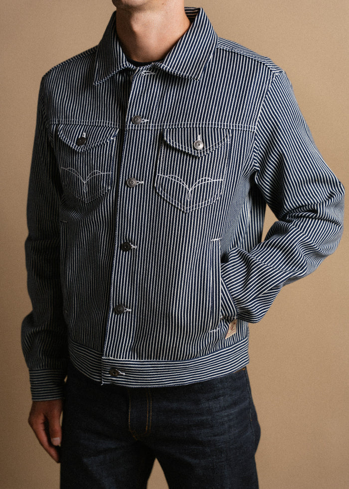 Man in a blue striped denim jacket with buttoned pockets stands against a beige background, showcasing casual fashion style.
