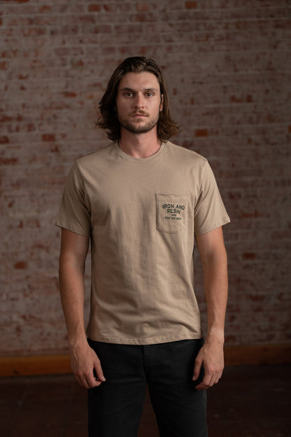 Man in beige "Iron and Resin" shirt stands against a brick wall, looking directly at the camera with a neutral expression.